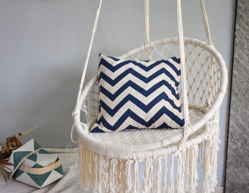 May include: A white macrame hanging chair with a blue and white chevron patterned pillow. The chair has a fringe around the bottom.