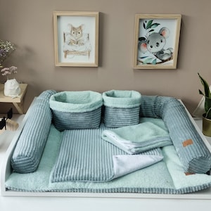 May include: A baby changing station with a soft, light blue corduroy surface, two matching baskets, and a bolster. Two framed illustrations of animals hang above. A wooden moose figurine and a small side table with flowers are also visible.