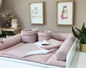 May include: A dusty pink waffle-textured baby changing mat set featuring a rectangular base with raised side bolsters, two matching cylindrical storage baskets, and a folded square cloth, all crafted from a soft, textured fabric material.