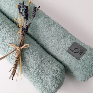 May include: Two rolled, pale teal bath towels. A small bundle of dried lavender and yellow flowers is tied with a brown ribbon and placed on top of one towel. A grey square patch with a black elephant logo is sewn on the other towel.
