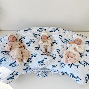 May include: A large, crescent-shaped nursing pillow featuring a light blue and white striped pattern with a repeating print of dark blue whales. Three infant dolls are positioned across the surface of the pillow, each wearing soft clothing.
