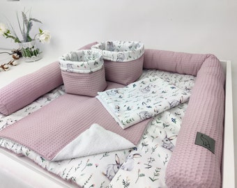 May include: A pink and white baby nest with a white sheet, two pink storage baskets, and a white towel. The sheet has a pattern of small white bunnies and green leaves.