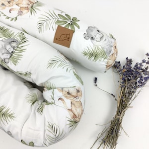 May include: Two white baby bolster pillows with a jungle animal print featuring lions, hippos, and palm leaves. A small brown leather tag is attached to one pillow. A bundle of dried lavender is next to the pillows.