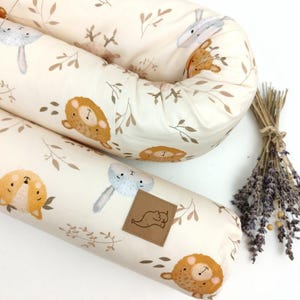 May include: A white and beige baby nest with a brown, white, and beige animal print. The nest is shaped like a long, curved cylinder and is filled with soft padding. The nest is ideal for newborns and infants.