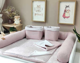 May include: A pink waffle-textured baby changing pad with a matching bolster, two square storage baskets, and a folded white and pink patterned blanket. The set features a soft fabric construction with a consistent light pink color palette throughout.