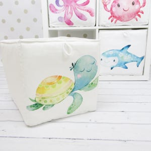 May include: A white fabric storage cube with a watercolour turtle illustration in green, yellow, and blue. Other cubes in the background display sea creature designs, including an octopus, crab, and shark. The cube has a loop handle.