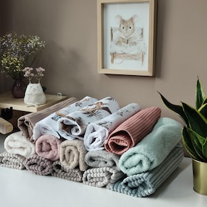Könnte beinhalten: Eine Sammlung gerollter Stoffhandtücher in verschiedenen Texturen und Farben, darunter Weiß, Beige, Altrosa, Salbeigrün und Grau, sowie gemusterte Tücher mit Illustrationen von Tieren und Blattwerk auf weißem Hintergrund.