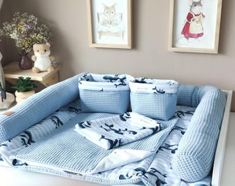 May include: A baby changing mat set featuring a light blue waffle-textured fabric with a white and dark blue whale pattern. The set includes two rectangular storage baskets, a padded mat with side bolsters, and a matching folded fabric cloth.