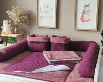 May include: A set of deep magenta waffle-textured nursery accessories including a long bolster cushion, two square storage baskets, and two folded rectangular cloths. The items feature a soft, plush interior lining and a consistent waffle-weave exterior.