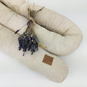 May include: A long, beige, plush, tubular pillow with a soft, textured surface. A small, square, brown leather-like patch with a line-art graphic of a sleeping animal is attached. A bundle of dried purple lavender stems is tied to the pillow.