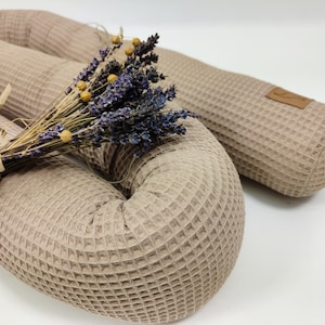 Op de afbeelding: Close-up van een beige wafelgeweven kussen met een bos gedroogde lavendel. Het kussen is cilindrisch en heeft een gestructureerd oppervlak. Een klein leren label is bevestigd aan het uiteinde van een kussen. De lavendel heeft paarse bloemen en groene stelen.