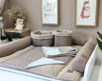 May include: A beige waffle-textured baby changing pad set featuring a rectangular base with raised side bolsters, two matching cylindrical storage baskets, and a folded square blanket. The fabric has a waffle weave pattern with a soft interior lining.