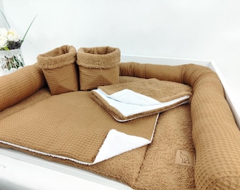 May include: A tan waffle-textured fabric set including a rectangular padded base with raised edges, two matching upright storage baskets, and a folded blanket. The items feature a white underside and a small square label with a line drawing of a bear.