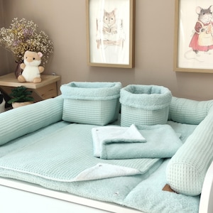 Può includere: Un set per il cambio neonato color verde menta in tessuto a nido d'ape, composto da una base rettangolare, due cuscini laterali cilindrici, due cestini portaoggetti e una coperta piegata, rifiniti con piccole etichette in similpelle marrone.