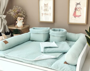 May include: A mint green baby changing pad set featuring a waffle-textured fabric, including a rectangular base, two cylindrical side bolsters, two storage baskets, and a folded blanket, all accented with small brown leather-like brand tags.