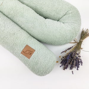 Op de afbeelding: Een lichtgroen, U-vormig kussen van badstof met een bruin leren label met een klein olifantontwerp. Het kussen ligt op een wit oppervlak naast een bos gedroogde lavendel.