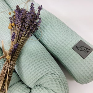 May include: Two light green waffle-textured cylindrical pillows with a small grey label featuring a black elephant illustration on one pillow. A bunch of dried lavender is placed on top of the pillows.