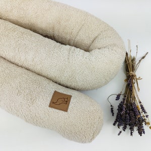 Op de afbeelding: Een beige, U-vormig kussen gemaakt van zacht, pluche stof. Het kussen heeft een bruin lederen label met een kleine, gestileerde dierillustratie.