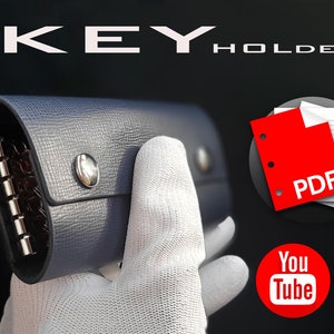 Puede incluir: Un llavero de cuero gris con un anillo de metal para llaves y un cierre de solapa. El llavero está siendo sostenido por una mano enguantada. La imagen también incluye un icono de PDF y un logotipo de YouTube.