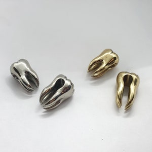 Könnte beinhalten: Vier silberne und goldene Zahn-Anhänger. Die Anhänger sind alle in verschiedenen Größen und Formen und haben eine glänzende Oberfläche.