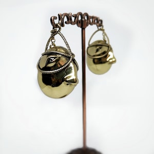 May include: A pair of gold-colored earrings with a face design. Each earring features a detailed face with a rope-like accent around the head. The earrings are displayed on a copper-colored stand against a white background.