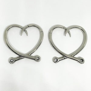May include: Two silver heart-shaped hooks with a curved point. The hooks are crossed at the bottom and have a small round loop at each end.