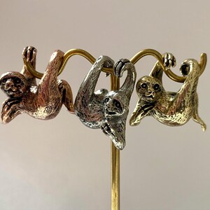 May include: Three metal sloth figurines in bronze, silver, and gold hang from a gold-colored bar. The sloths are detailed with textured fur and facial features. The figurines are attached to a vertical gold-colored stand.