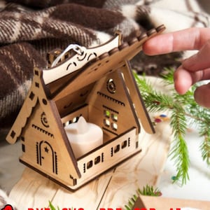 Könnte beinhalten: Ein Kerzenhalter aus Holz in Form eines Hauses mit einem aufklappbaren Dach. Der Halter ist aus lasergeschnittenem Holz gefertigt und hat ein detailliertes Design. Der Kerzenhalter steht auf einer Holzoberfläche.