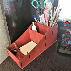 Może przedstawiać: Drewniany organizer biurkowy w kolorze czerwonym z wieloma przegródkami. Organizer jest wypełniony różnymi artykułami biurowymi, w tym nożyczkami, długopisami, pędzlami i butelką kleju.