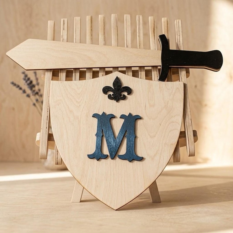 A light wood shield with a dark blue letter M and a fleur-de-lis, resting on a stand with a wooden sword behind it against a light wood panel background.