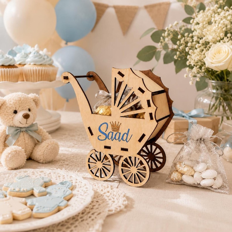 A party scene featuring the wooden baby stroller toy on a table surrounded by a teddy bear, blue and white cupcakes, cookies, balloons, and a vase of flowers.