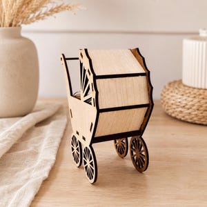 A rear view of the laser-cut wooden baby stroller toy showing its rounded hood and small spoked wheels on a light wood surface next to a white vase and cloth.