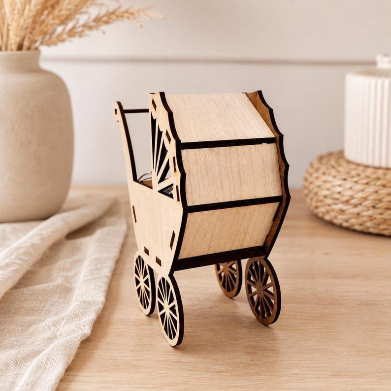 A rear view of the laser-cut wooden baby stroller toy showing its rounded hood and small spoked wheels on a light wood surface next to a white vase and cloth.