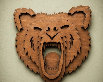 May include: A layered wooden wall art piece shaped like a roaring bear head. The design features a dark brown stained finish with intricate cutouts defining the fur texture, eyes, nose, and an open mouth revealing sharp teeth in a layered relief.