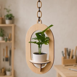 May include: A light-colored wooden oval hanging planter frame with a central shelf holding a white ceramic pot containing a green leafy plant. The frame is suspended by a vertical chain consisting of alternating light and dark wooden oval links.