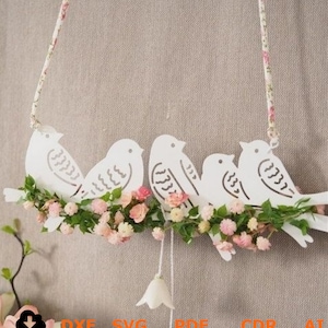 Könnte beinhalten: Weißer Metallausschnitt von fünf Vögeln, die auf einem Zweig mit rosa und weißen Blüten sitzen. Die Vögel sind in einer Reihe angeordnet und haben ein schlichtes Design. Der Zweig ist mit grünen Blättern und Blüten verziert.