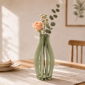 May include: A light green wooden vase with a vertical slat design and a clear glass insert. The vase holds a single peach-colored rose, a sprig of small white flowers, and a stem of eucalyptus leaves, creating a floral arrangement.