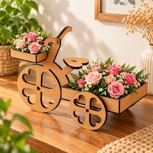 Può includere: Un portafiori in legno color marrone chiaro a forma di bicicletta con due cassette rettangolari. Le ruote e il sellino presentano intagli a forma di cuore. Il supporto è riempito con rose rosa, piccoli fiori bianchi e fogliame verde.