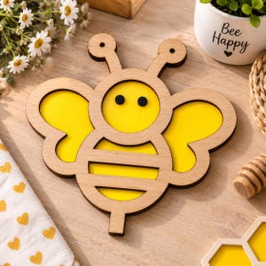 Könnte beinhalten: Ein dekorativer Holzausschnitt in Bienenform mit einem Rahmen aus hellem Naturholz, leuchtend gelben Einsätzen für Flügel und Körper, zwei schwarzen runden Augen und zwei Antennen mit runden Spitzen, konzipiert als flaches Wandobjekt.