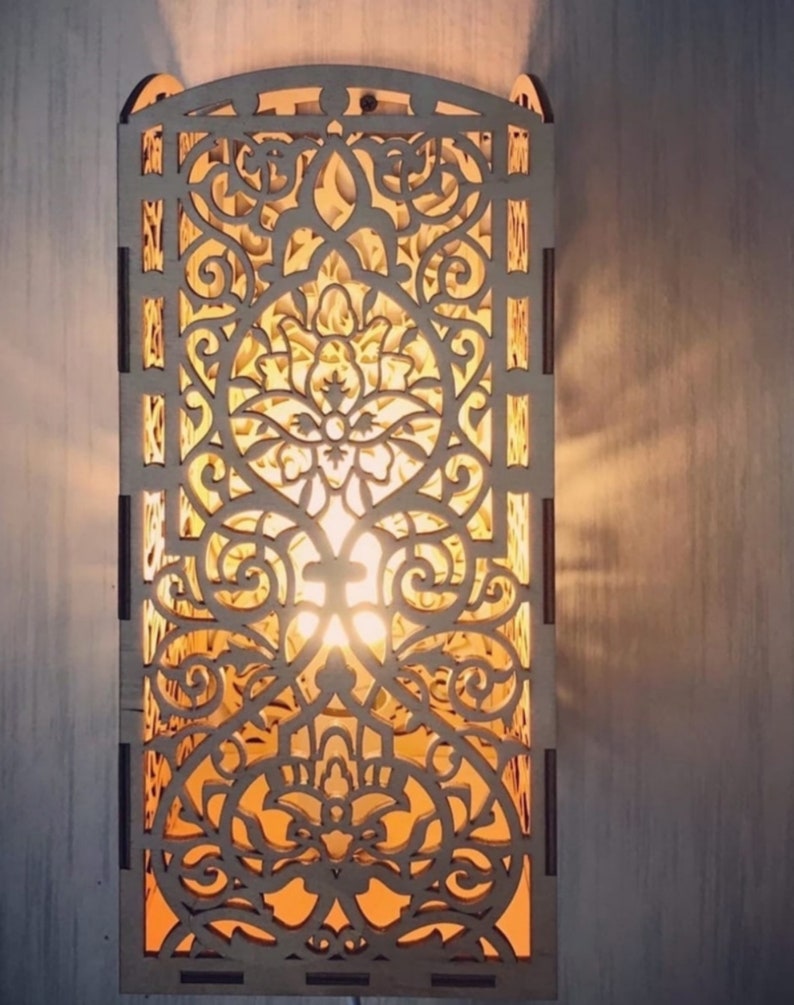 May include: A wooden wall sconce with an intricate, laser-cut floral design. The sconce is rectangular with a curved top and emits a warm, inviting light. The light shines through the detailed patterns, creating a decorative and artistic effect.
