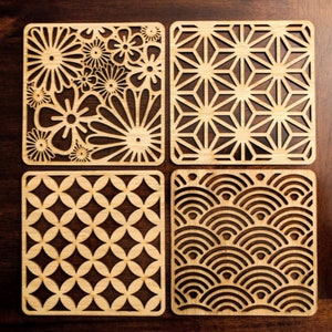 May include: Four square wooden coasters with intricate laser-cut designs. The coasters feature a floral pattern, a geometric pattern, a circular pattern, and a wave pattern.
