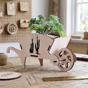 May include: A light-colored wooden wheelbarrow planter featuring a black laser-cut cactus silhouette on the side. The planter holds a green succulent plant with small, rounded leaves. The structure is assembled with visible slots and a single wheel.