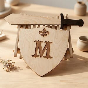 A light wood shield featuring a brown letter M and a fleur-de-lis, placed on a table stand with a wooden sword and small dried flowers nearby.