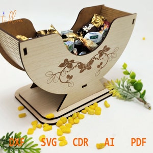 Puede incluir: Un cuenco de madera para dulces con forma de cuna, con un diseño floral y una base. El cuenco está lleno de chocolates y dulces surtidos.