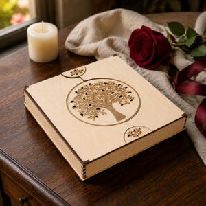 May include: A square light-colored wooden box featuring a laser-cut circular design of a tree on its lid. The box has dark brown edges with visible interlocking joints at the corners and two small circular cutouts above and below the center.