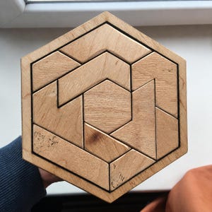 May include: A hexagonal wooden puzzle consisting of several geometric pieces fitted together within a matching hexagonal frame. The light-colored wood features a natural grain pattern, and the individual segments form a complex, interlocking design.