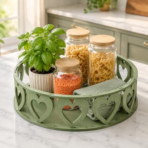 May include: A round sage green wooden tray with heart-shaped cutouts along the rim. Inside the tray sits a green basil plant in a ribbed white pot, three clear glass jars with bamboo lids containing pasta and lentils, and a folded green cloth.