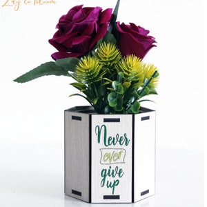 May include: A wooden hexagonal planter with the text "Never ever give up" in green and yellow lettering. The planter is filled with artificial flowers, including three burgundy roses and green foliage.