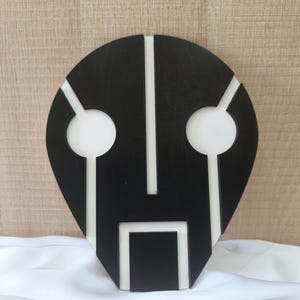 May include: Black and white geometric mask with a stylized face design. The mask is made of wood and has a smooth finish.