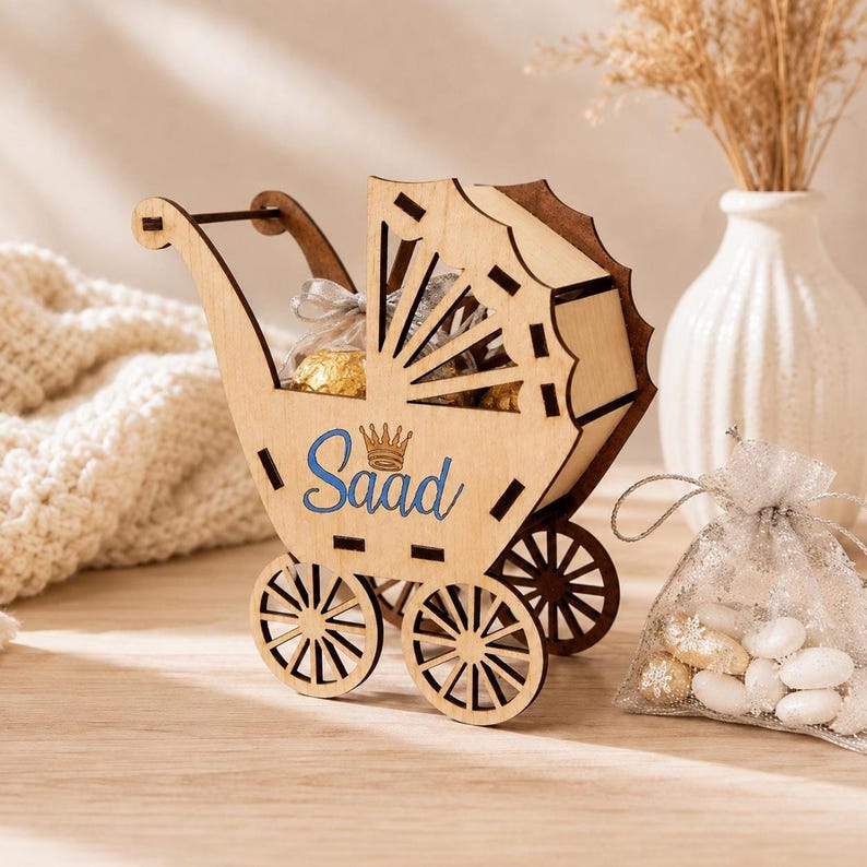 The wooden toy baby stroller with the name Saad on the side stands on a wooden table next to a sheer organza drawstring bag filled with white and gold candies.